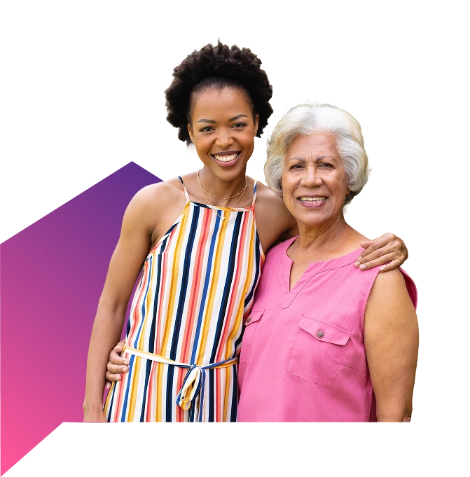 A younger woman in a colorful striped dress is smiling and standing with her arm around an older woman in a pink top.