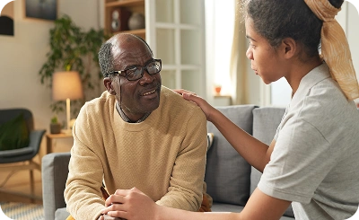 An older man in a tan sweater sits on a couch, engaged in conversation with a young woman. She comforts him with a hand on his shoulder, conveying empathy.