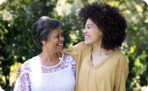 Two women stand outside, smiling warmly at each other. One wears a white patterned blouse, the other a yellow top. Lush greenery is in the background.