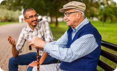 A young man and an elderly man sit on a park bench, engaged in a friendly conversation. The elderly man holds a cane, and both appear relaxed and smiling.