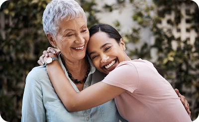 A joyful, smiling young woman in a pink shirt embraces an elderly woman in a light blue shirt outdoors, conveying warmth and affection amidst greenery.