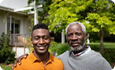 A young man in an orange shirt smiles with his arm around an older man in a gray sweater. They're outdoors, with trees and a house in the background, conveying warmth.