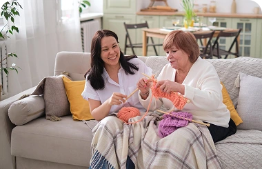 Caregiver helping an elderly woman knit on a sofa in a cozy living room