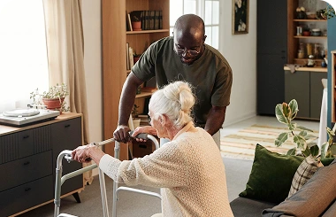 Caregiver assisting an elderly woman with a walker while standing up at home.
