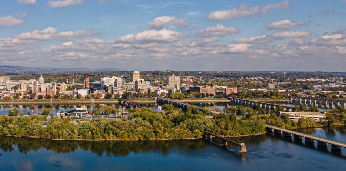 Aerial view of a city skyline with buildings along a river. A bridge crosses the water near a lush green island. The sky is dotted with fluffy clouds.