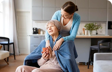 Caregiver comforting an elderly woman with a blanket on a sofa at home