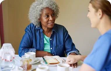 Older woman with curly gray hair in a denim shirt chatting over breakfast with a caregiver in blue scrubs.