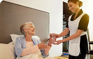 Smiling caregiver hands a glass of water to an elderly woman sitting up in bed.