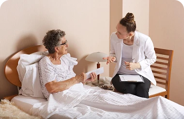 Female healthcare professional in a white coat discussing medication with an older woman sitting up in bed.