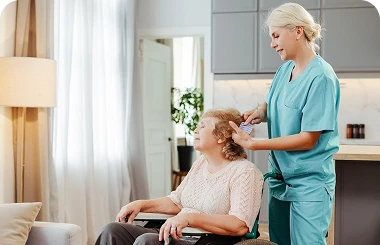 Female caregiver in teal scrubs gently brushing an elderly woman’s hair while she sits in a wheelchair in a cozy home setting.